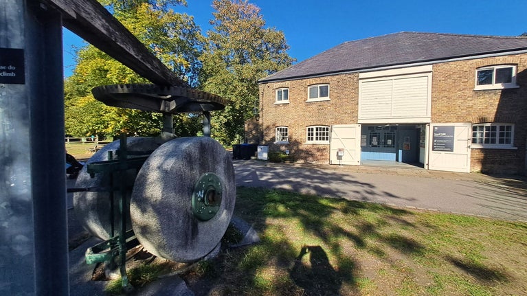 arge stone grinding wheel and overhead wooden drive mechanism in the foreground outside the Snuff Mill at Morden Hall Park. The brick mill building stands behind with blue entrance doors, surrounded by trees and dappled sunlight under a clear blue sky.